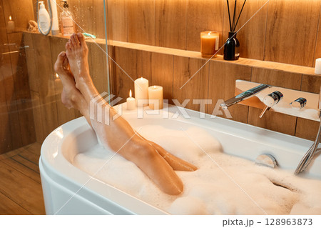 Female legs in bathtub filled with foam and warm lighting. Woman relaxing in bathroom with candles. Concept of wellness, relaxation, and self care 128963873