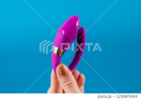 A woman holds a curved sex toy in lubrication on a blue background. Vaginally clitoral vibrator. A woman holds a curved sex toy in lubrication on a blue background. Vaginally clitoral vibrator. 128965684