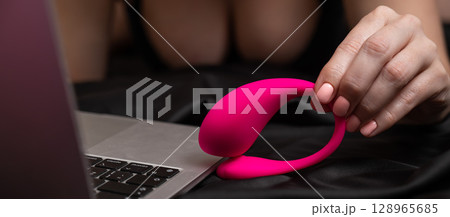 A woman is lying in bed holding a curved vibrator and looking at a laptop. A woman is lying in bed holding a curved vibrator and looking at a laptop. 128965685