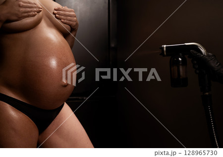 Faceless pregnant woman getting an instant tan. The master applies bronzer with a spray. 128965730