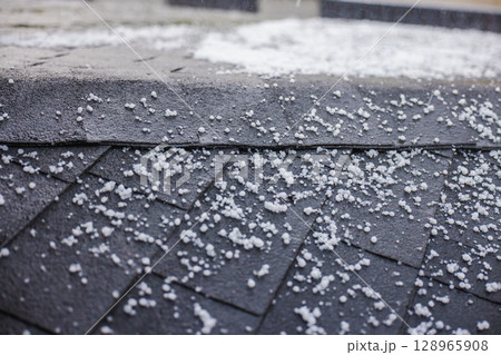 Close-up of small hailstones scattered on a black roof with shingles during a hailstorm. Close-up of small hailstones scattered on a black roof with shingles during a hailstorm. 128965908