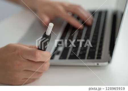 A woman smokes an electronic cigarette while working on a laptop. Tobacco heating system. 128965919