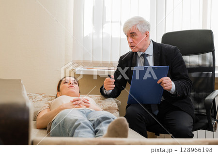 Caucasian woman lies on a couch at a reception with a psychotherapist. An elderly man works as a psychiatrist Caucasian woman lies on a couch at a reception with a psychotherapist. An elderly man works as a psychiatrist 128966198
