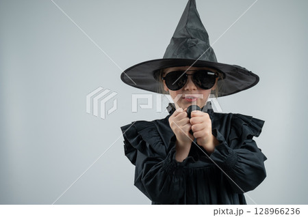 Portrait of a little Caucasian girl in a witch costume inflates a black balloon on a white background. Portrait of a little Caucasian girl in a witch costume inflates a black balloon on a white background. 128966236