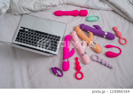 Collection of sex toys on a white sheet. Various vibrators and a laptop on the bed. Collection of sex toys on a white sheet. Various vibrators and a laptop on the bed. 128966384