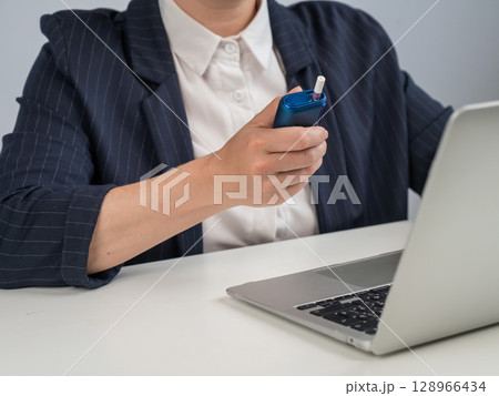 Business woman smoking an electronic cigarette while working on a laptop. Tobacco heating system. Business woman smoking an electronic cigarette while working on a laptop. Tobacco heating system. 128966434