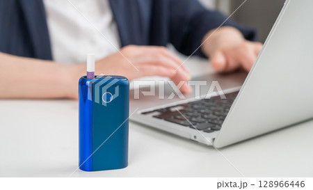 Business woman smoking an electronic cigarette while working on a laptop. Tobacco heating system in the foreground. 128966446
