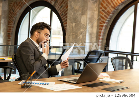 Young businessman scrutinizing document and looking concentrated Young businessman scrutinizing document and looking concentrated 128966604