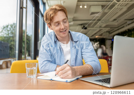Ginger young man sitting at the table in the cafe and making notes Ginger young man sitting at the table in the cafe and making notes 128966611