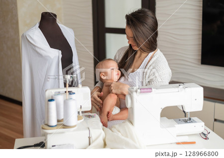 A young woman sews in the kitchen and holds a small child. Mom teaches her little son to sew on a sewing machine. Self-isolation homework 128966800