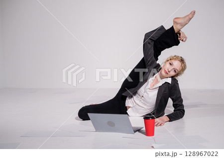 Barefoot ballerina dressed in a business suit poses for a laptop and drinks coffee. Flexible woman works at the computer 128967022