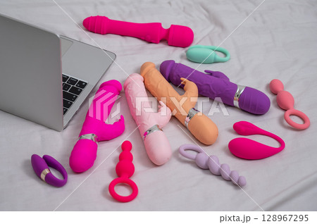Collection of sex toys on a white sheet. Various vibrators and a laptop on the bed.  128967295