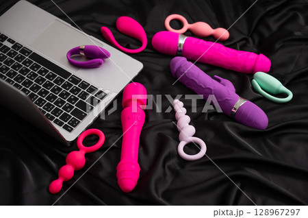 Collection of sex toys on a black sheet. Top view of various vibrators and a laptop. Collection of sex toys on a black sheet. Top view of various vibrators and a laptop. 128967297