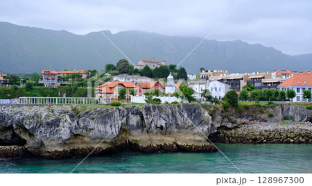 Picturesque coastal town in Northern Spain, featuring traditional houses with red roofs perched atop dramatic cliffs overlooking a turquoise bay. Llane,s, Asturias Picturesque coastal town in Northern Spain, featuring traditional houses with red roofs perched atop dramatic cliffs overlooking a turquoise bay. Llane,s, Asturias 128967300