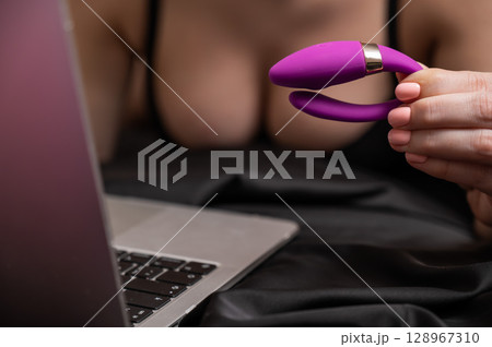 A woman is lying in bed holding a curved vibrator and looking at a laptop. A woman is lying in bed holding a curved vibrator and looking at a laptop. 128967310