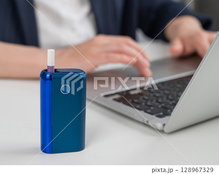 Business woman smoking an electronic cigarette while working on a laptop. Tobacco heating system in the foreground. Business woman smoking an electronic cigarette while working on a laptop. Tobacco heating system in the foreground. 128967329