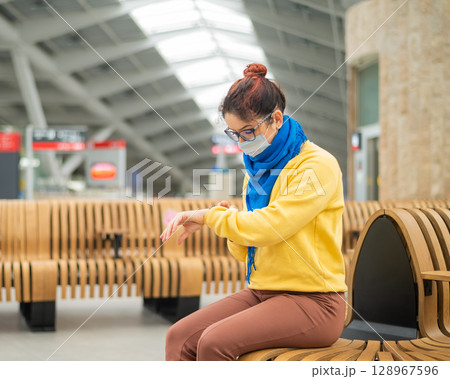 Caucasian woman in mask uses smart watch on bench in public place. Caucasian woman in mask uses smart watch on bench in public place. 128967596