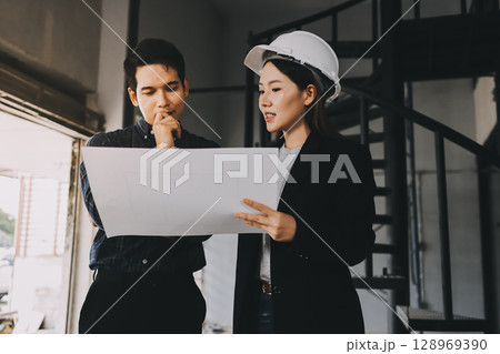 Construction manager and engineer dressed in orange work vests and hard helmets explore construction documentation on the building site near the steel frames 128969390