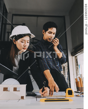 Construction manager and engineer dressed in orange work vests and hard helmets explore construction documentation on the building site near the steel frames 128969394