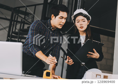 Construction manager and engineer dressed in orange work vests and hard helmets explore construction documentation on the building site near the steel frames 128969400
