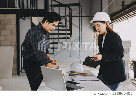 Construction manager and engineer dressed in orange work vests and hard helmets explore construction documentation on the building site near the steel frames 128969405