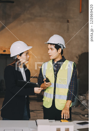 Construction manager and engineer dressed in orange work vests and hard helmets explore construction documentation on the building site near the steel frames 128969420