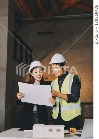 Construction manager and engineer dressed in orange work vests and hard helmets explore construction documentation on the building site near the steel frames 128969426