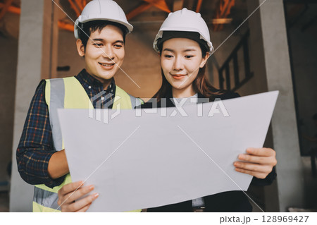 Construction manager and engineer dressed in orange work vests and hard helmets explore construction documentation on the building site near the steel frames Construction manager and engineer dressed in orange work vests and hard helmets explore construction documentation on the building site near the steel frames 128969427