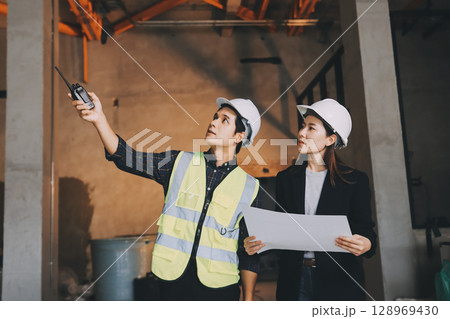 Construction manager and engineer dressed in orange work vests and hard helmets explore construction documentation on the building site near the steel frames 128969430