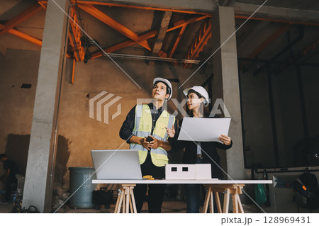 Construction manager and engineer dressed in orange work vests and hard helmets explore construction documentation on the building site near the steel frames Construction manager and engineer dressed in orange work vests and hard helmets explore construction documentation on the building site near the steel frames 128969431