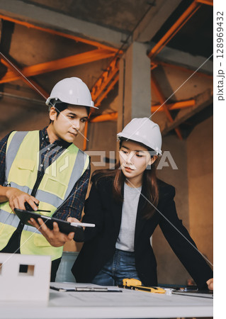 Construction manager and engineer dressed in orange work vests and hard helmets explore construction documentation on the building site near the steel frames Construction manager and engineer dressed in orange work vests and hard helmets explore construction documentation on the building site near the steel frames 128969432