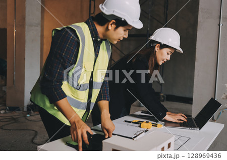 Construction manager and engineer dressed in orange work vests and hard helmets explore construction documentation on the building site near the steel frames Construction manager and engineer dressed in orange work vests and hard helmets explore construction documentation on the building site near the steel frames 128969436