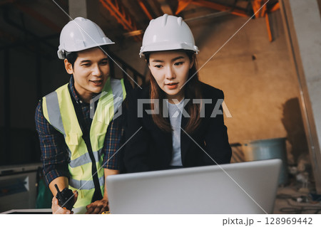 Construction manager and engineer dressed in orange work vests and hard helmets explore construction documentation on the building site near the steel frames 128969442