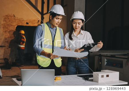 Construction manager and engineer dressed in orange work vests and hard helmets explore construction documentation on the building site near the steel frames Construction manager and engineer dressed in orange work vests and hard helmets explore construction documentation on the building site near the steel frames 128969443