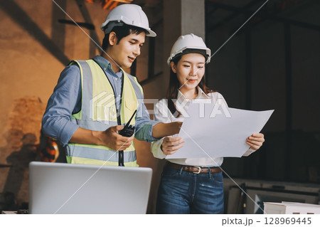 Construction manager and engineer dressed in orange work vests and hard helmets explore construction documentation on the building site near the steel frames Construction manager and engineer dressed in orange work vests and hard helmets explore construction documentation on the building site near the steel frames 128969445