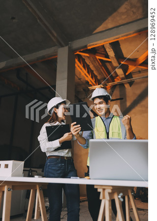 Construction manager and engineer dressed in orange work vests and hard helmets explore construction documentation on the building site near the steel frames 128969452