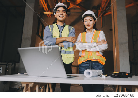 Construction manager and engineer dressed in orange work vests and hard helmets explore construction documentation on the building site near the steel frames Construction manager and engineer dressed in orange work vests and hard helmets explore construction documentation on the building site near the steel frames 128969469
