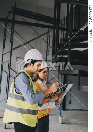 Construction manager and engineer dressed in orange work vests and hard helmets explore construction documentation on the building site near the steel frames Construction manager and engineer dressed in orange work vests and hard helmets explore construction documentation on the building site near the steel frames 128969474