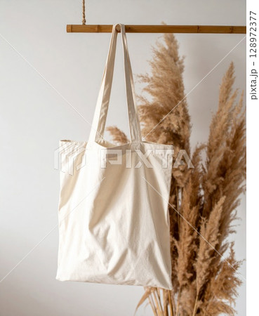 Beige Canvas Tote Bag Hanging Against White Wall with Pampas Grass Decoration Indoors 128972377