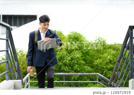 Businessman walking up stairs with laptop and coffee, looking at his watch 128972939