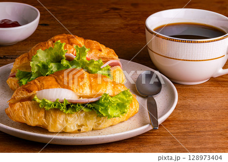 Croissant ham cheese with green vegetable and slice onion in brown spotted ceramic plate with a cup of black coffee. 128973404