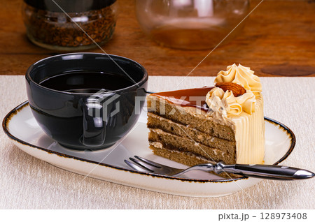 Side view of a cup of black coffee and a slice of coffee layered cake garnished with cream and almond. 128973408