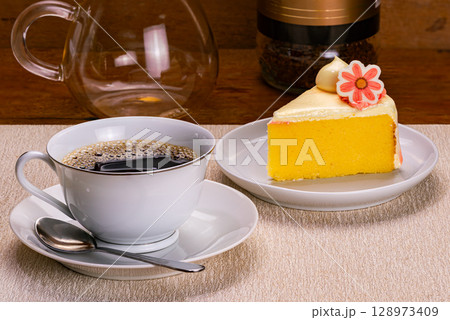Side view of a cup of black coffee in white ceramic cup with a metal spoon on white ceramic saucer and slice of vanilla sponge cake. Side view of a cup of black coffee in white ceramic cup with a metal spoon on white ceramic saucer and slice of vanilla sponge cake. 128973409