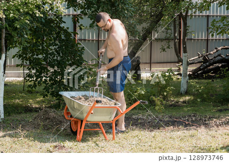 Shirtless Man Gardening with Wheelbarrow in Backyard: Sunny Outdoor Scene, Green Foliage, Orange Tool, Rural Landscape, Realistic Photography 128973746