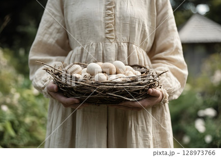 A woman in a light-colored dress holds a woven basket filled with fresh eggs. The background features a lush garden and a chicken coop, symbolizing slow living and outdoor harvest. 128973766