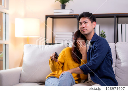 Young asian couple watching a scary or thrilling TV show in living room at home with emotion shock. Young asian couple watching a scary or thrilling TV show in living room at home with emotion shock. 128975607