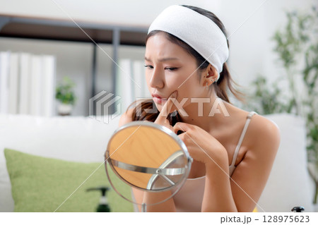 Young asian woman looking mirror with stress on facial sitting on sofa in living room at home. Young asian woman looking mirror with stress on facial sitting on sofa in living room at home. 128975623