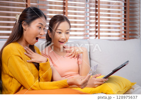 Two young asian woman sitting on sofa using tablet computer shopping online with credit card buying. 128975637