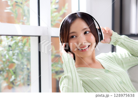 Happy young asian woman wearing headphones listening music while sitting on sofa with fun. 128975638