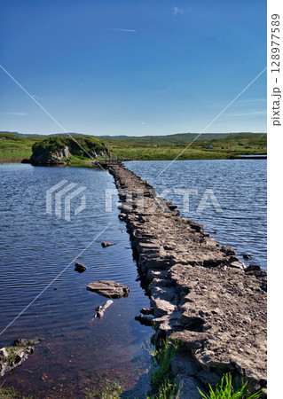 Scenic view in Ireland with a stone path leading across a tranquil lake towards a small island under a bright blue sky. Scenic view in Ireland with a stone path leading across a tranquil lake towards a small island under a bright blue sky. 128977589